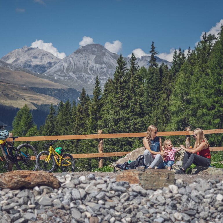 Zwei Frauen und ein Mädchen sitzen am Ufer eines Bergsees, daneben steht ein grosses und ein kleines Bike. | © Davos Klosters Mountains