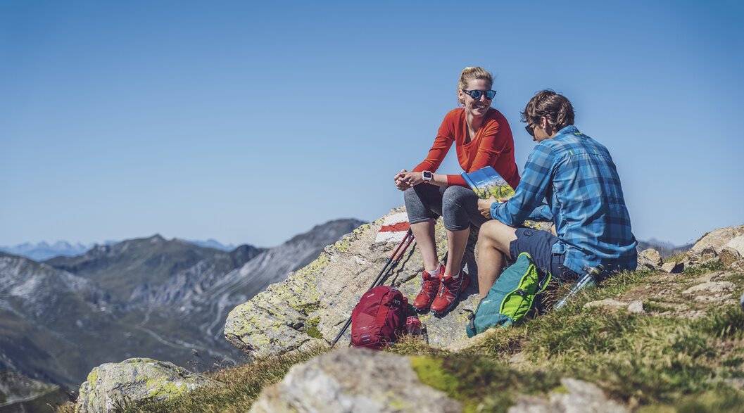 Ein Paar sitzt auf einem Stein auf dem Gipfel,  während der Rast studiert der Mann die Wanderkarte und die Frau geniesst die Sonne.  | © Davos Klosters Mountains