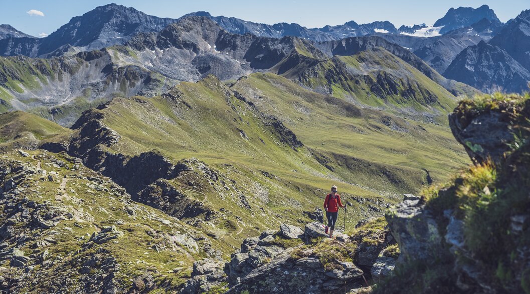 Eine in rot gekleidete Wanderin steigt mit ihren Stöcken den steinigen Grat hinauf, hinter ihr öffnet sich der Blick in das Sertig Tal.  | © Davos Klosters Mountains
