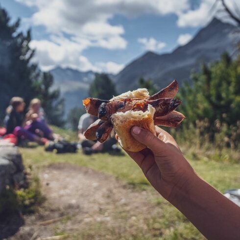 Ein Kind hält sein Brötchen mit der eingeklemmten Cervelat in die Höhe, dahinter die Grillstelle. | © Davos Klosters Mountains